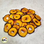 اسلایس‌های حلقه‌ای آلو کالیفرنیا خشک با پوست تیره مایل به مشکی و گوشت زرد طلایی که روی سطحی سفید قرار دارند.