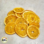 اسلایس‌های پرتقال تامسون خشک با رنگ نارنجی روشن در بسته بندی شفاف و طلایی آسان میوه خشک که تعدادی از آن‌ها بیرون بسته ریخته شده است.