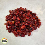انبوهی از دانه‌های کرن بری خشک با رنگ قرمز تیره که روی سطحی خاکستری و روشن قرار گرفته‌اند.