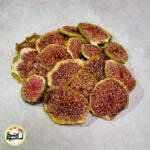 اسلایس‌های حلقه‌ای انجیر سبز خشک با پوست سبز و مغز قرمز رنگ که روی سطحی خاکستری قرار گرفته‌اند.