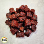 انبوهی از تکه‌های مکعبی و تیره رنگ حبه آلو فشرده با رنگ قرمز تیره که روی سطحی خاکستری و روشن قرار گرفته‌اند.
