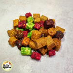 انبوهی از مخلوط حبه‌های میوه‌ای ایرانی با رنگ‌های متنوع قهوه‌ای، قرمز، سبز و نارنجی که روی سطحی خاکستری و روشن قرار گرفته‌اند.